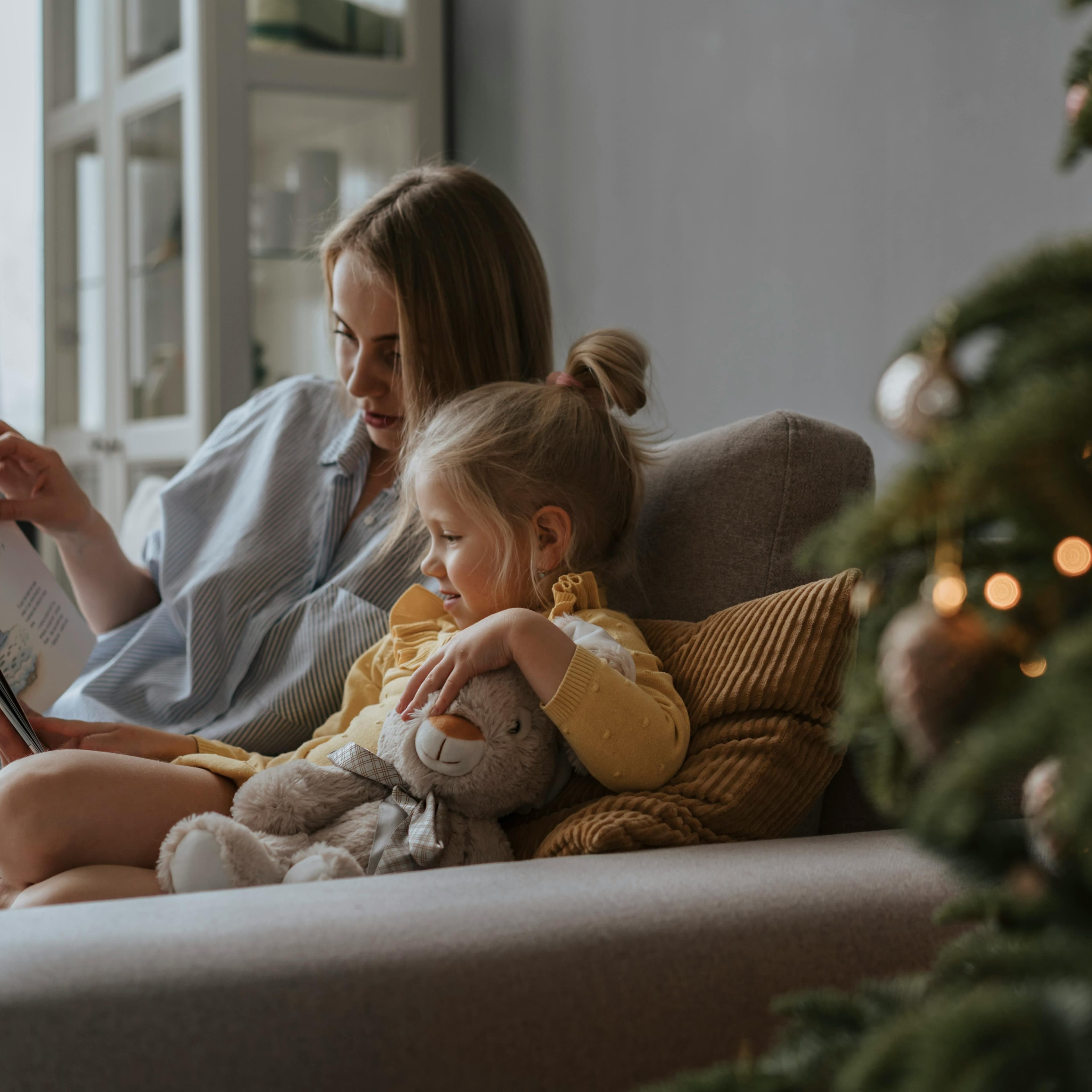 Mother and young daughter sitting on a couch by a Christmas tree and reading together