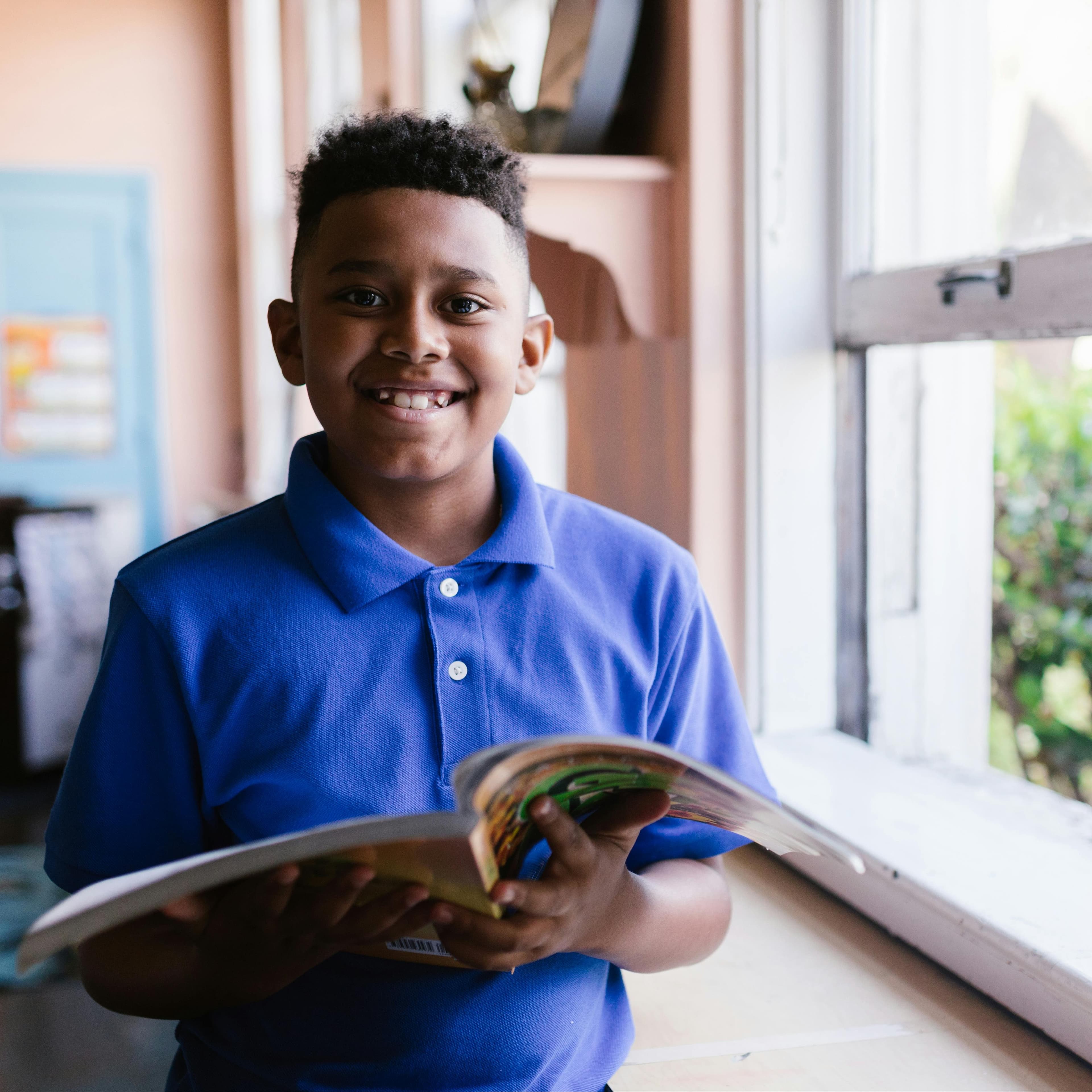 Smiling boy holding a book in a classroom by an open window