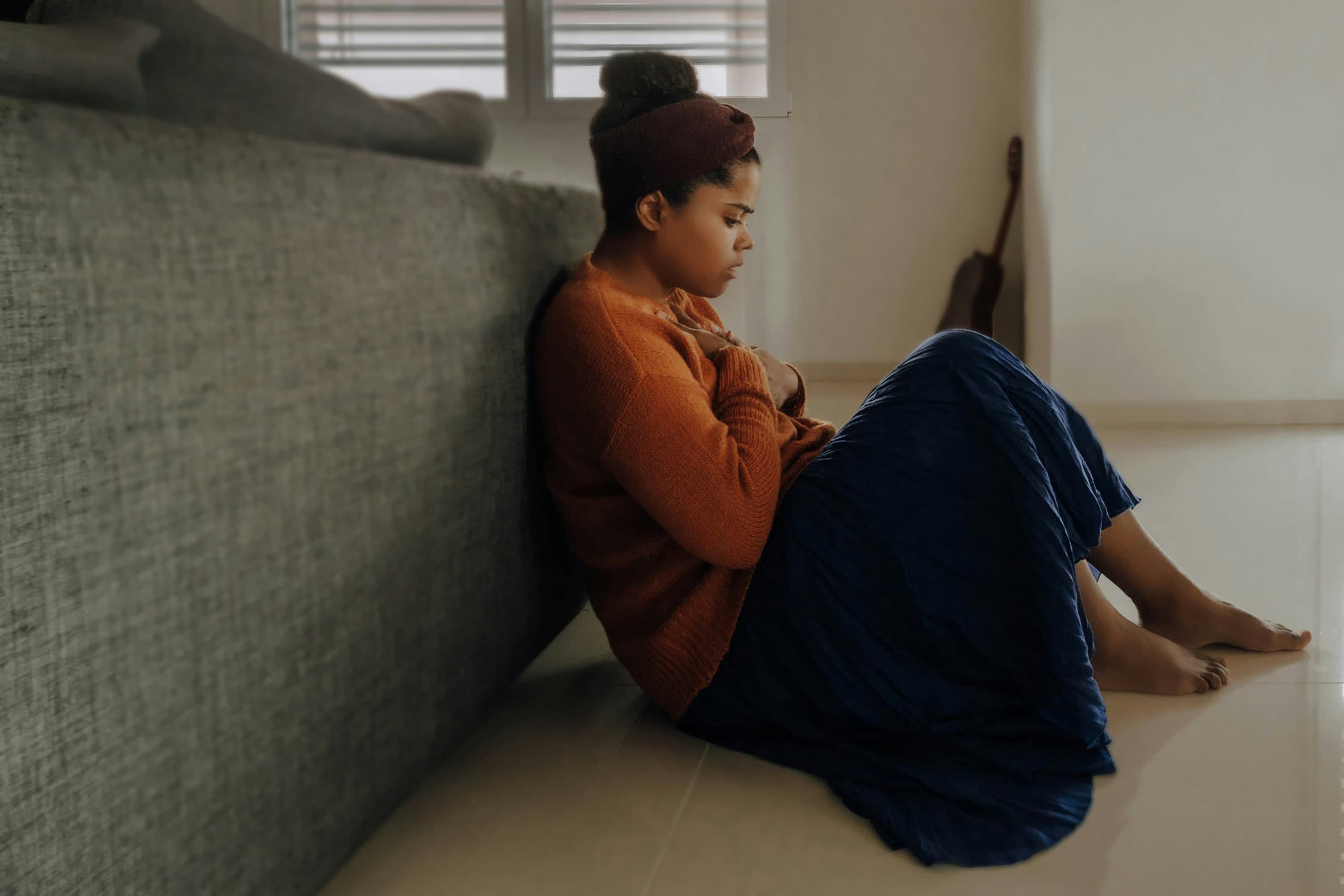 Young teen girl sitting on the floor leaning against the back of the couch, hands on her chest, breathing through her mouth and looking down