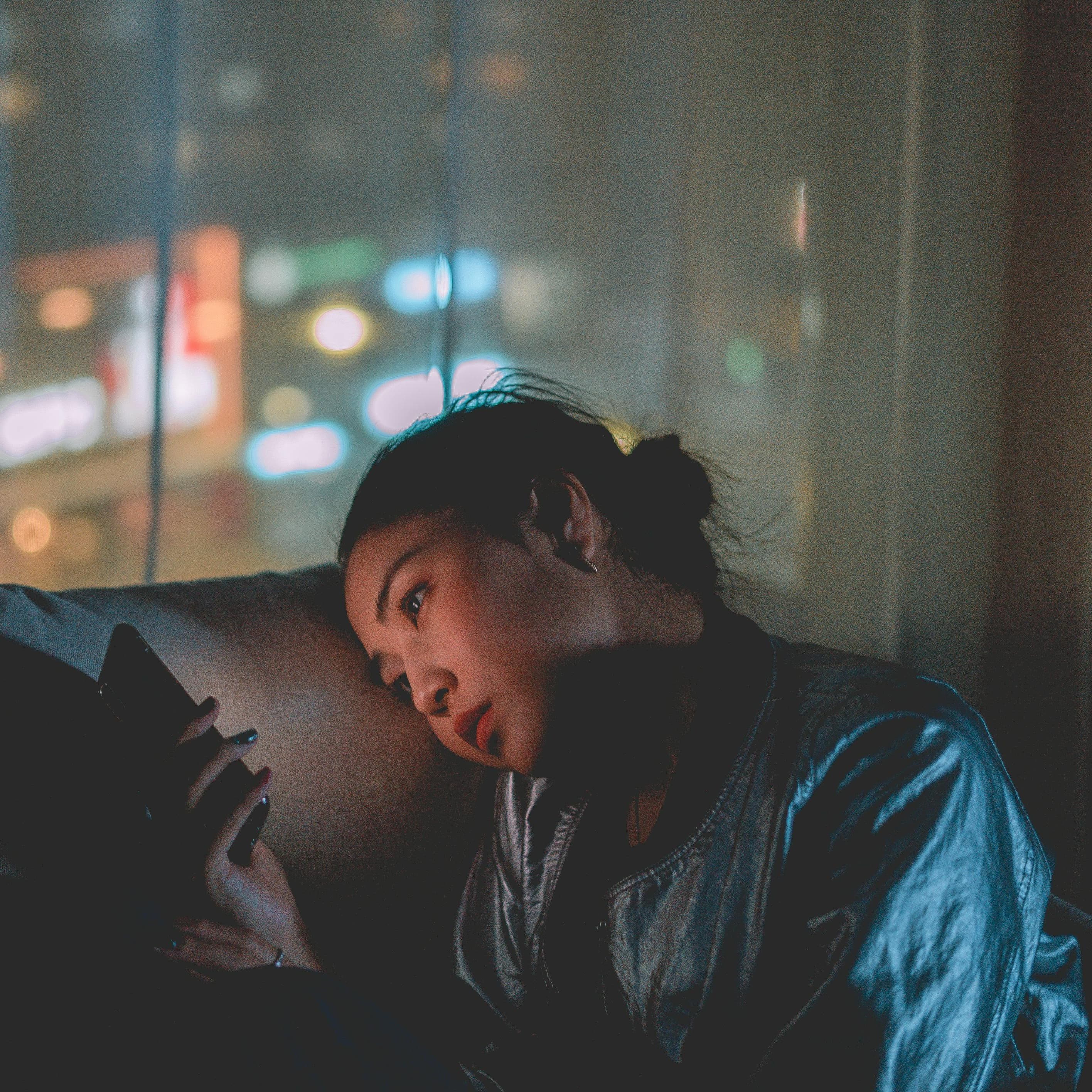 Young woman sits on a couch in a dark room looking at her phone with the city in the background out the windows