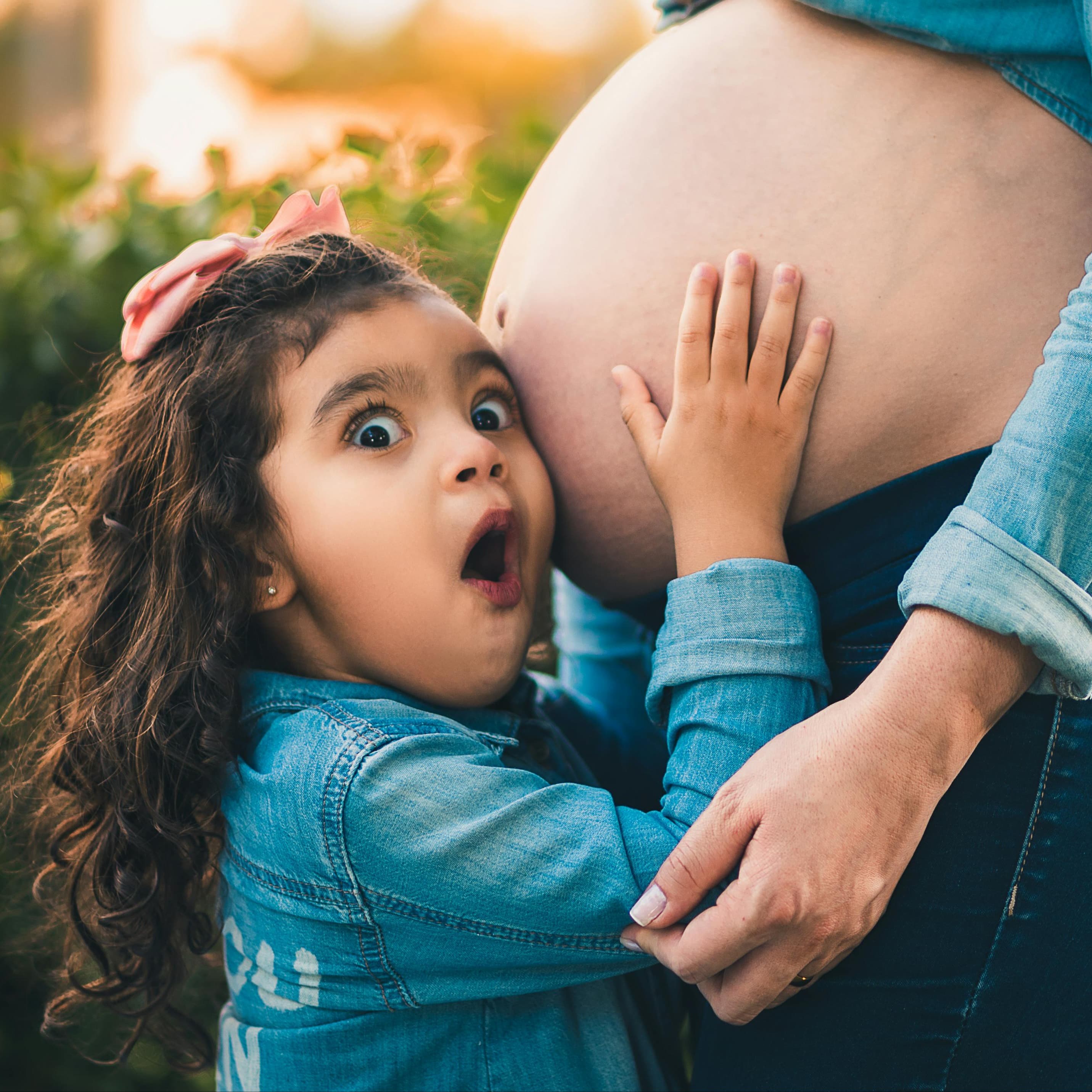 Young girl is listening to her mother's pregnant belly and has a surprised look on her face