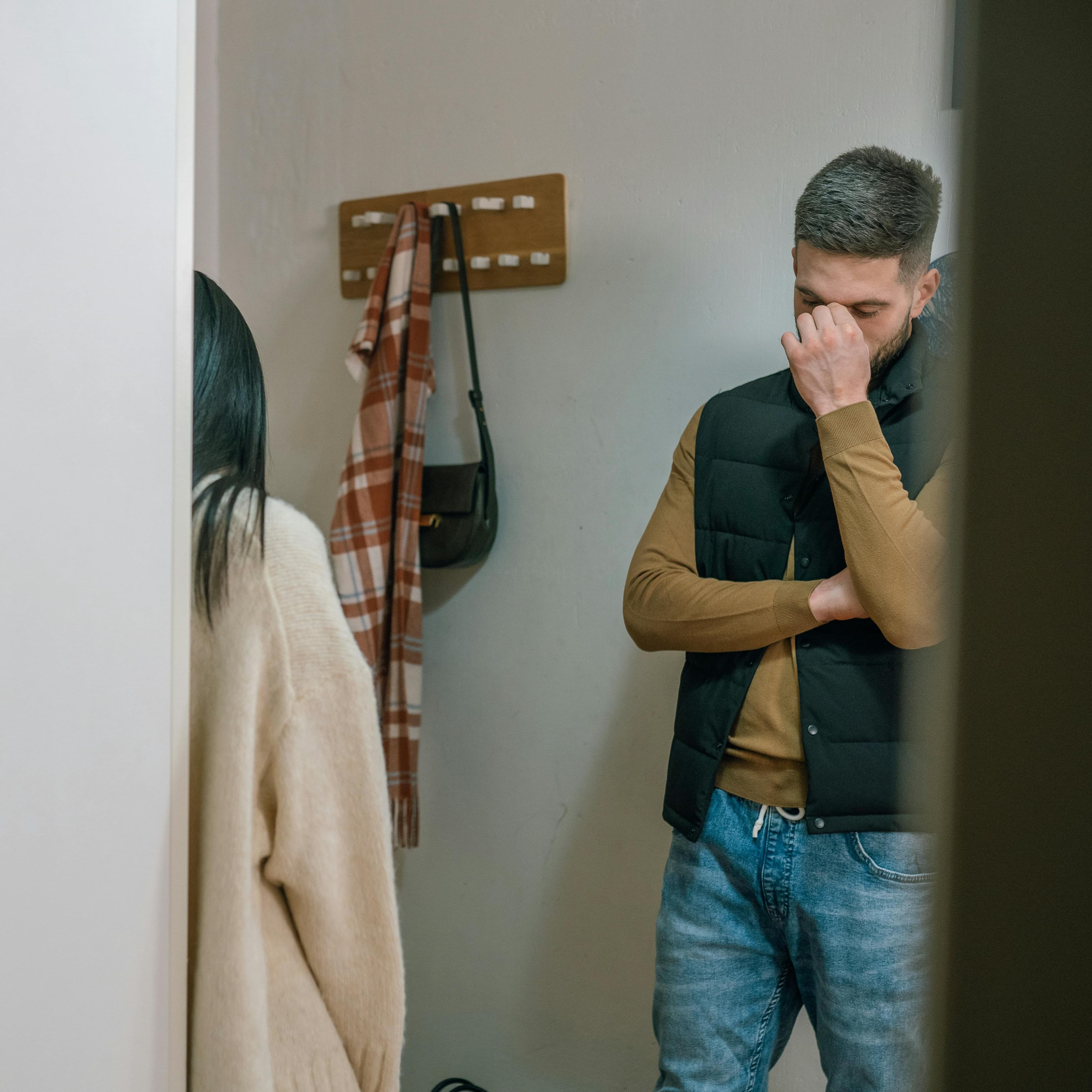 Man and woman standing in hallway across from each other not looking at each other, man has arm crossed over his chest and other hand up at the bridge of his nose