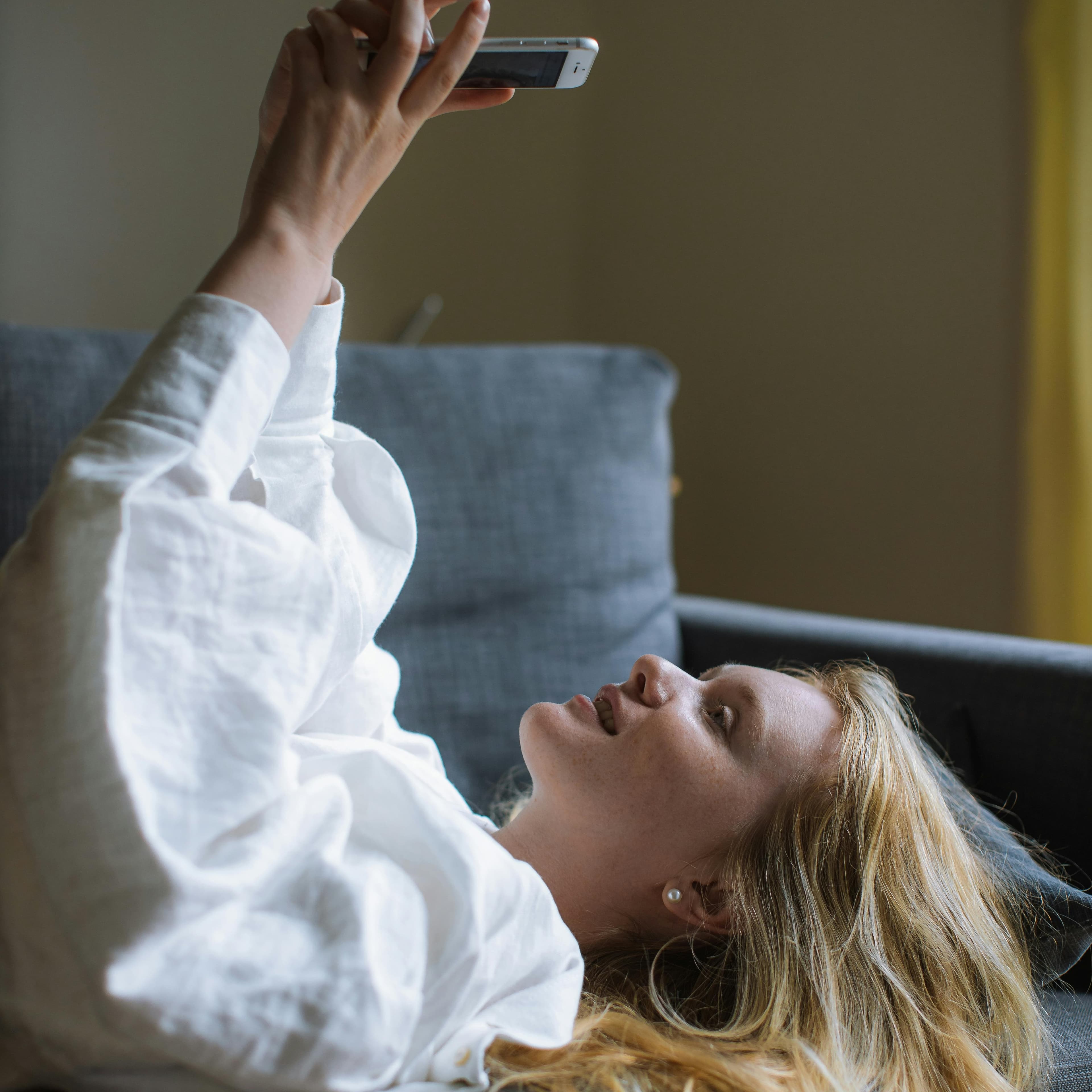 Young woman laying on her back on the couch with her phone overhead, looking at it with a smile
