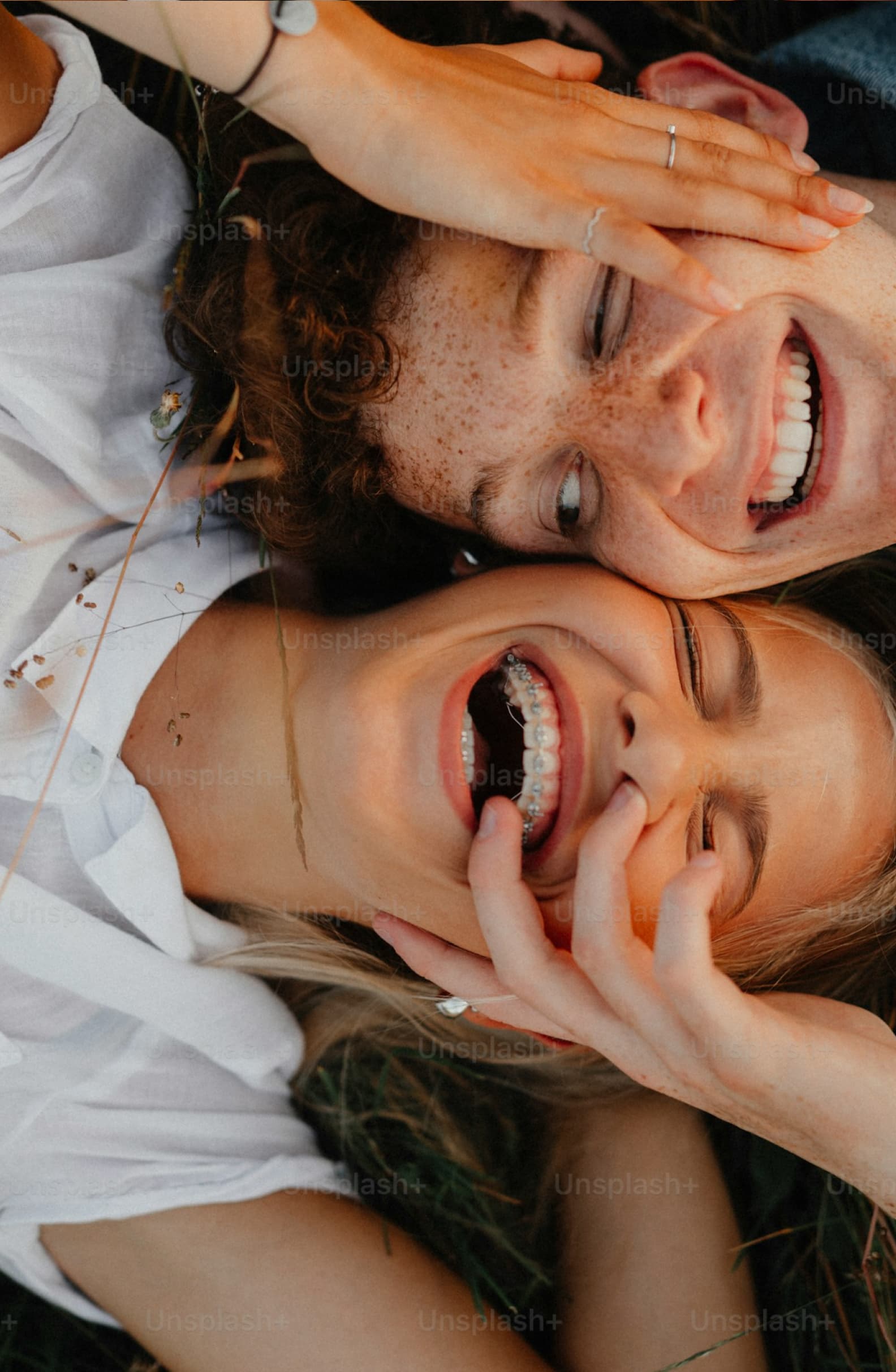 Candid overhead shot of two people lying down wearing white shirts, both showing bright smiles with dental braces, displaying genuine joy and friendship
